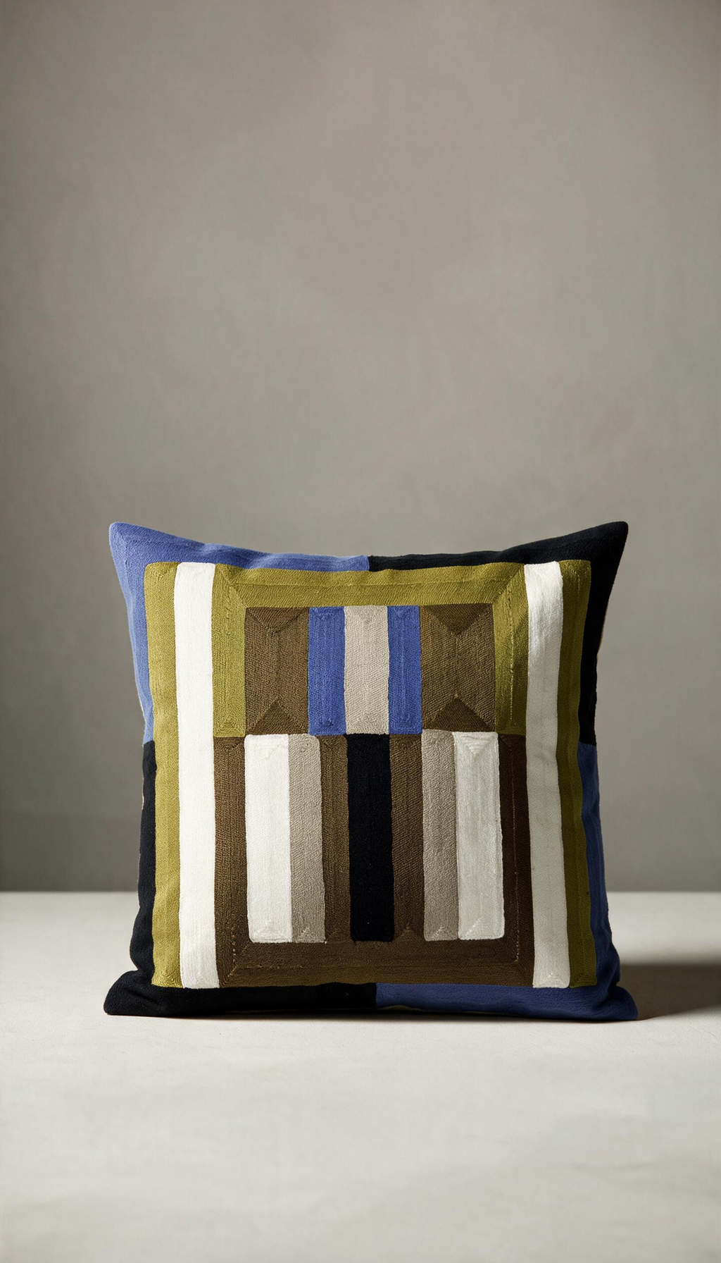Cojín modelo Jalisco de 45x45cm, con patrón geométrico bordado a rayas y bloques combinando tonos tierra, oliva, negro, crema y detalles en azul eléctrico.