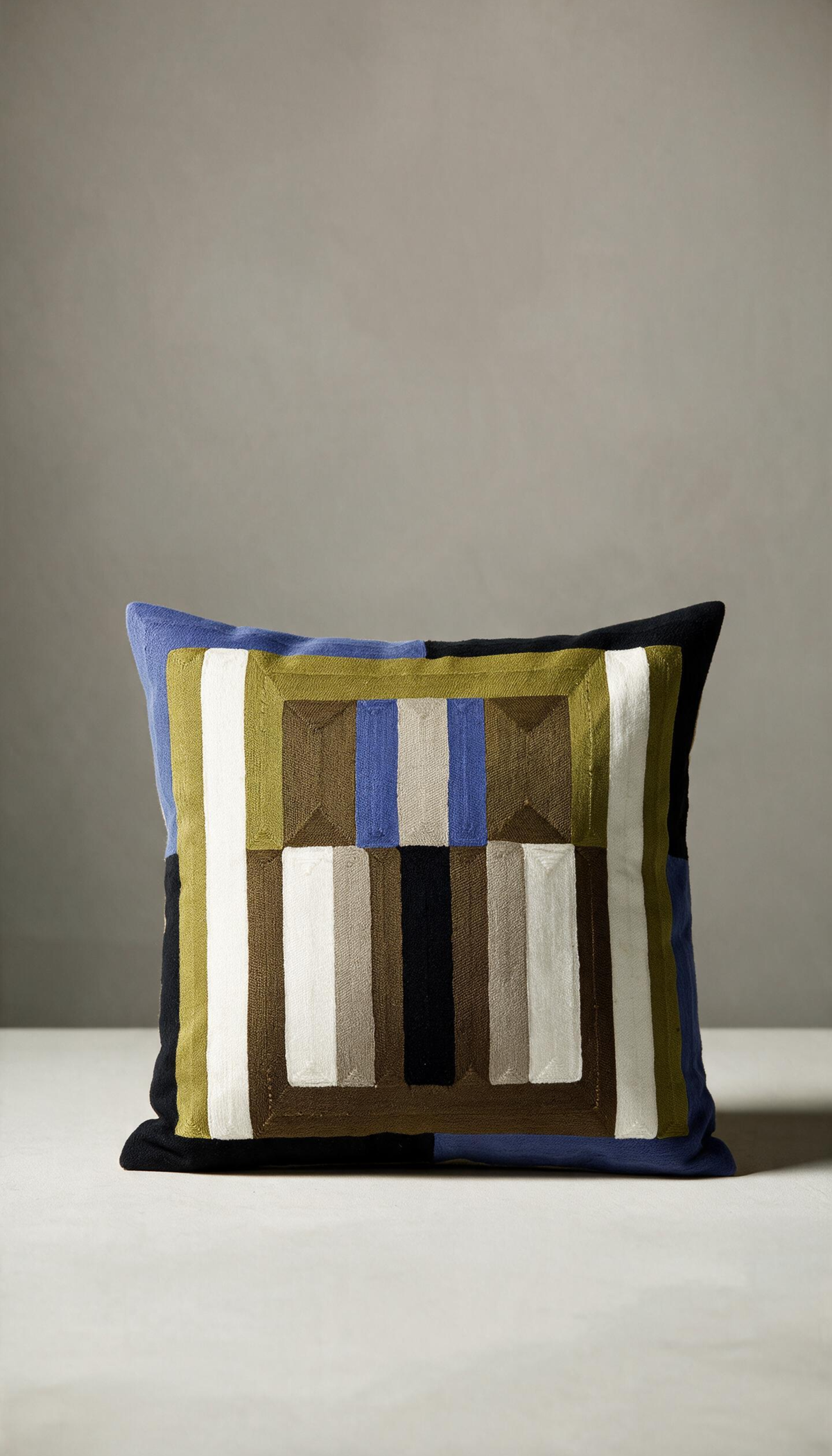 Cojín modelo Jalisco de 45x45cm, con patrón geométrico bordado a rayas y bloques combinando tonos tierra, oliva, negro, crema y detalles en azul eléctrico.
