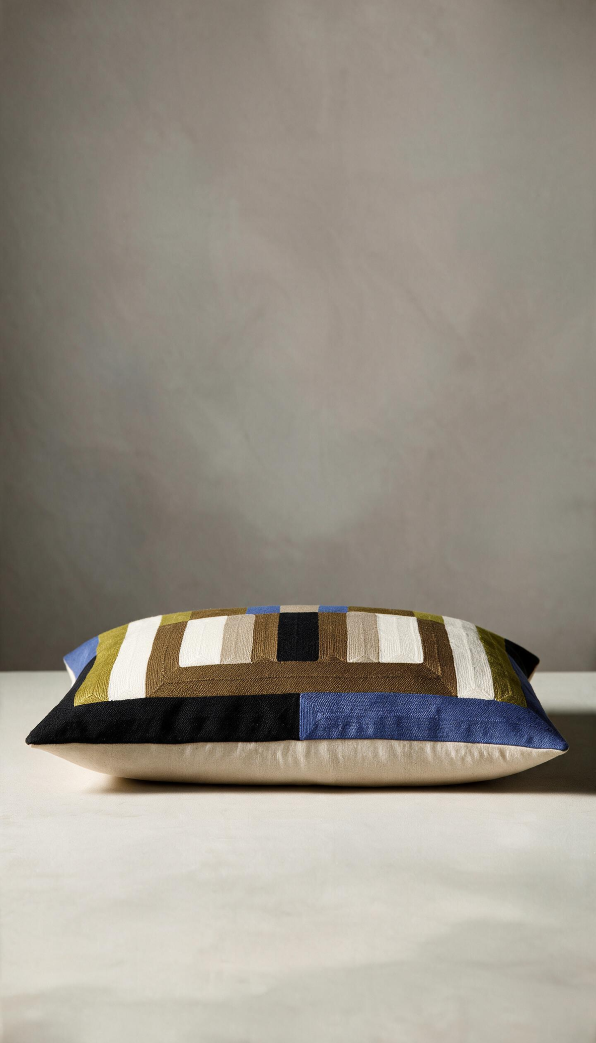 Cojín modelo Jalisco de 45x45cm, con patrón geométrico bordado a rayas y bloques combinando tonos tierra, oliva, negro, crema y detalles en azul eléctrico.
