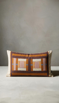 Cojín rectangular modelo Brezo con diseño de estampado geométrico en bloques combinando colores tierra, terracota, marrón y rosa sobre un fondo claro natural.