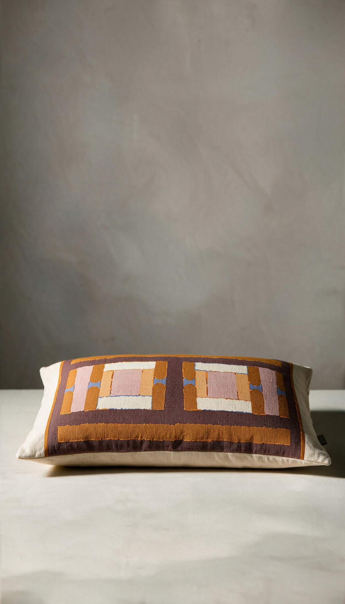 Cojín rectangular modelo Brezo con diseño de estampado geométrico en bloques combinando colores tierra, terracota, marrón y rosa sobre un fondo claro natural.