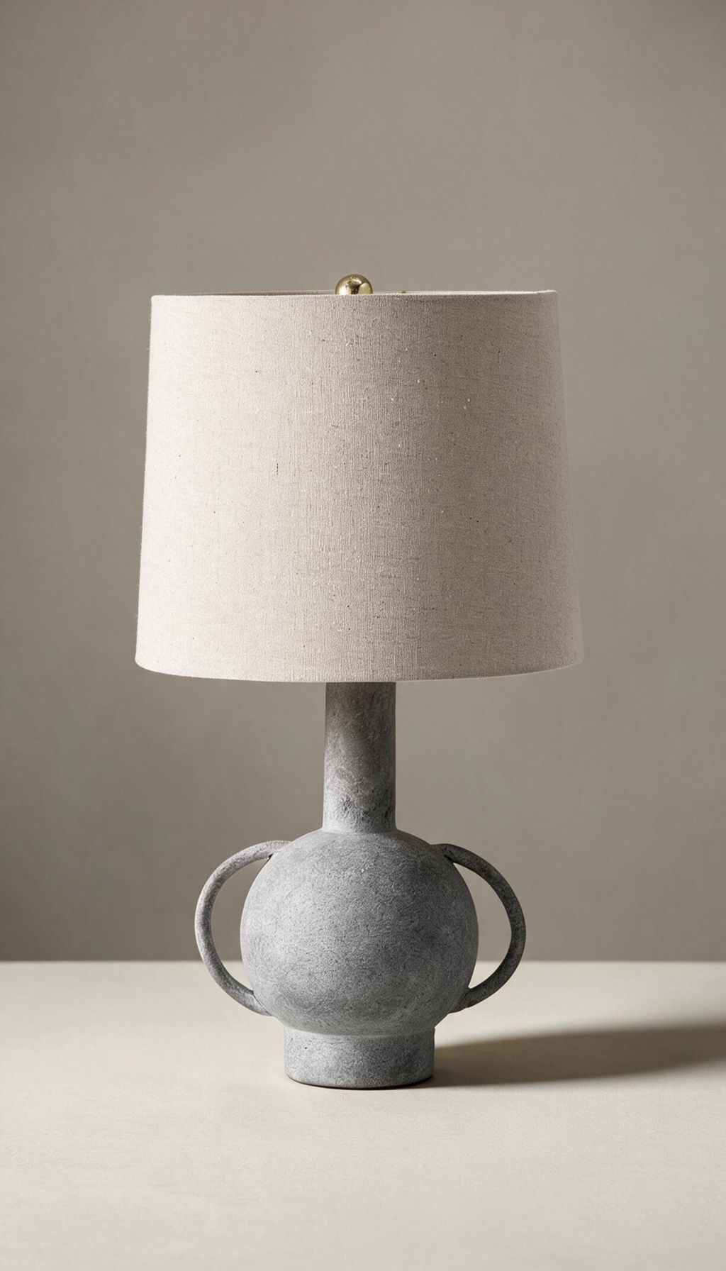 Lámpara de mesa modelo Umbral con base escultural tipo ánfora en gris texturizado efecto piedra, y pantalla cilíndrica de tela de lino color natural.
