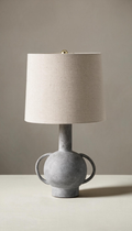 Lámpara de mesa modelo Umbral con base escultural tipo ánfora en gris texturizado efecto piedra, y pantalla cilíndrica de tela de lino color natural.