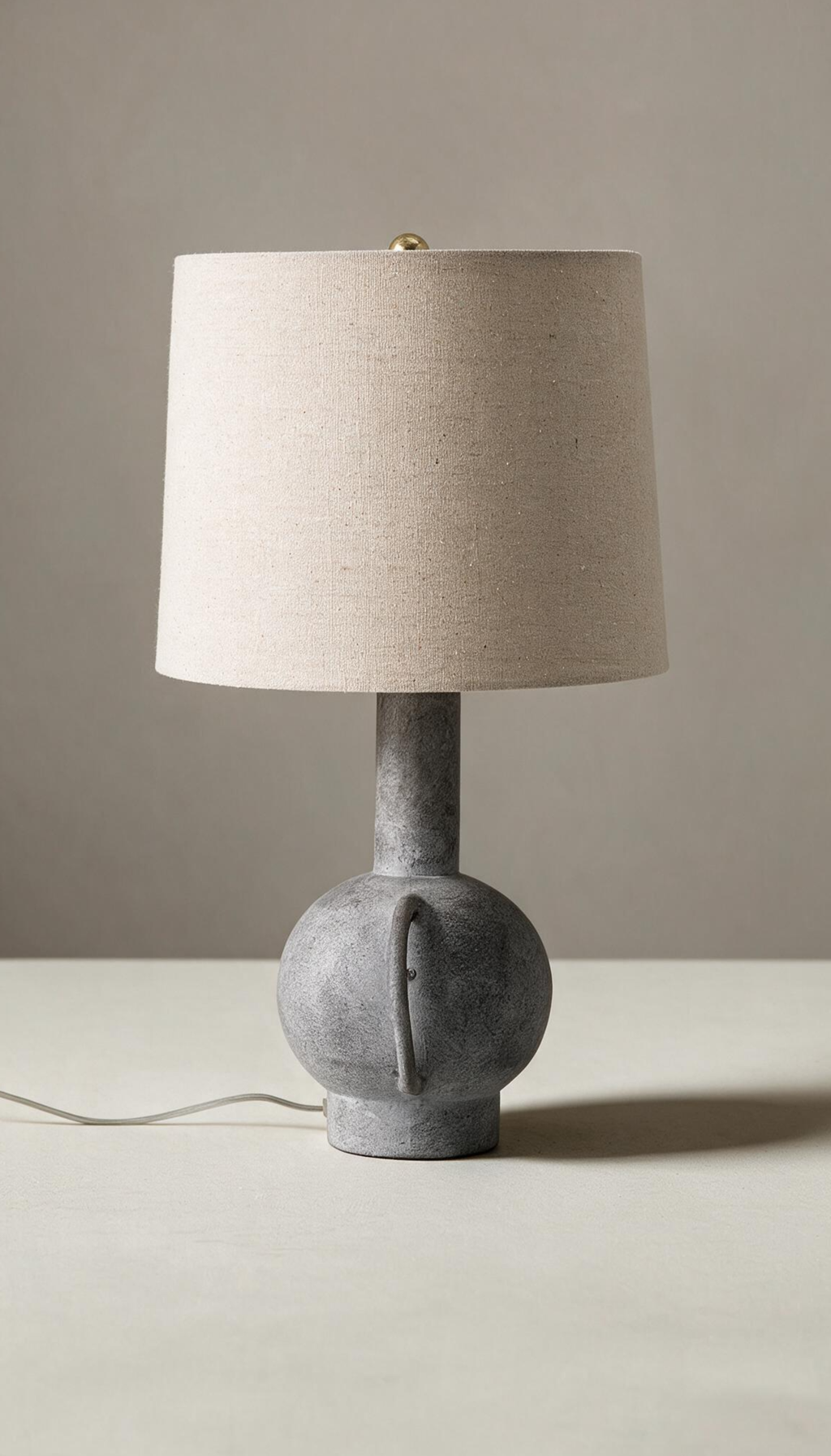 Lámpara de mesa modelo Umbral con base escultural tipo ánfora en gris texturizado efecto piedra, y pantalla cilíndrica de tela de lino color natural.