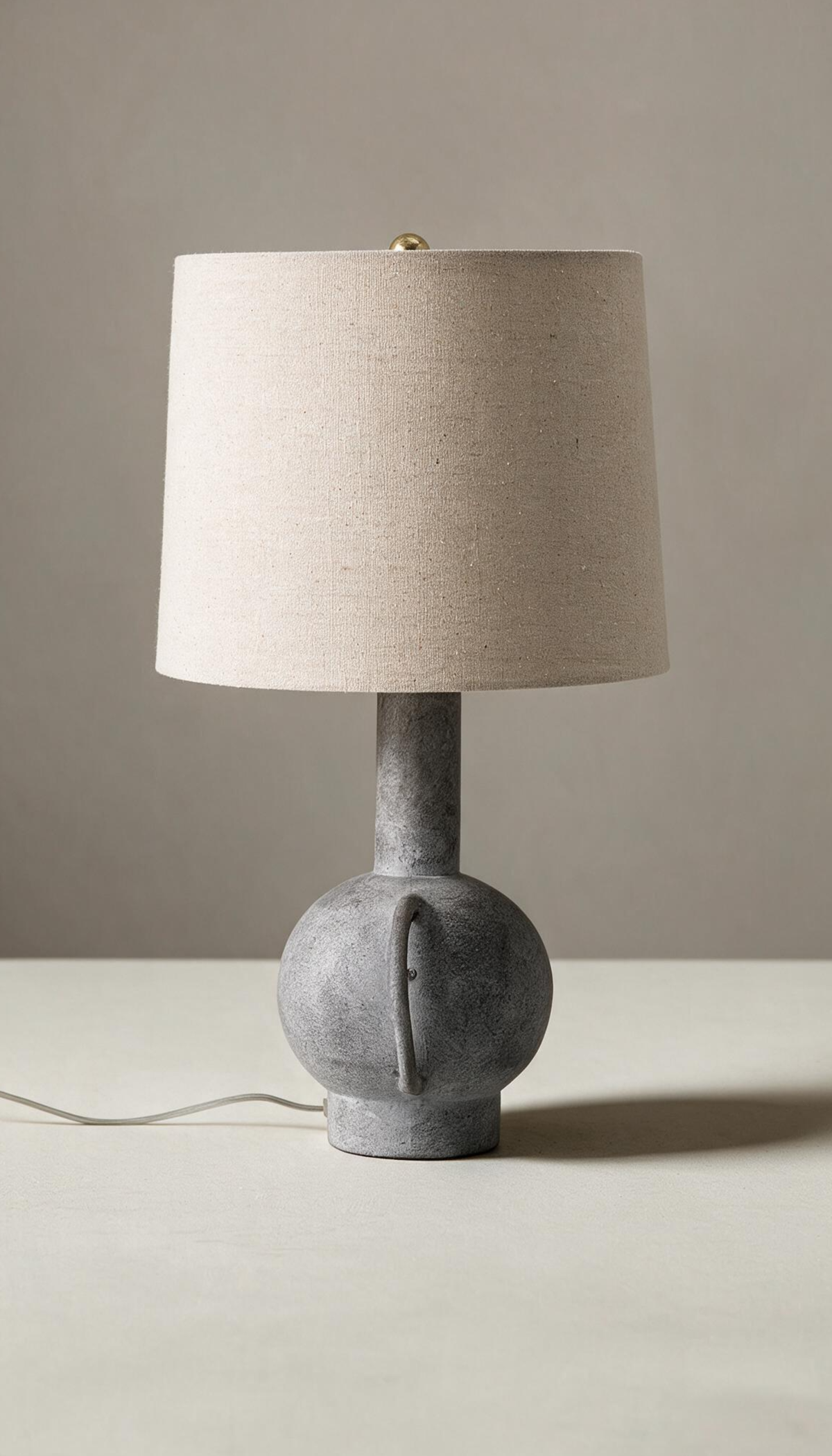 Lámpara de mesa modelo Umbral con base escultural tipo ánfora en gris texturizado efecto piedra, y pantalla cilíndrica de tela de lino color natural.