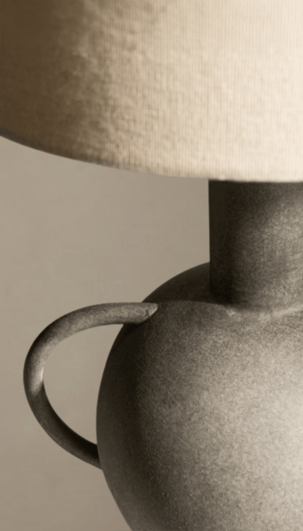 Lámpara de mesa modelo Umbral con base escultural tipo ánfora en gris texturizado efecto piedra, y pantalla cilíndrica de tela de lino color natural.