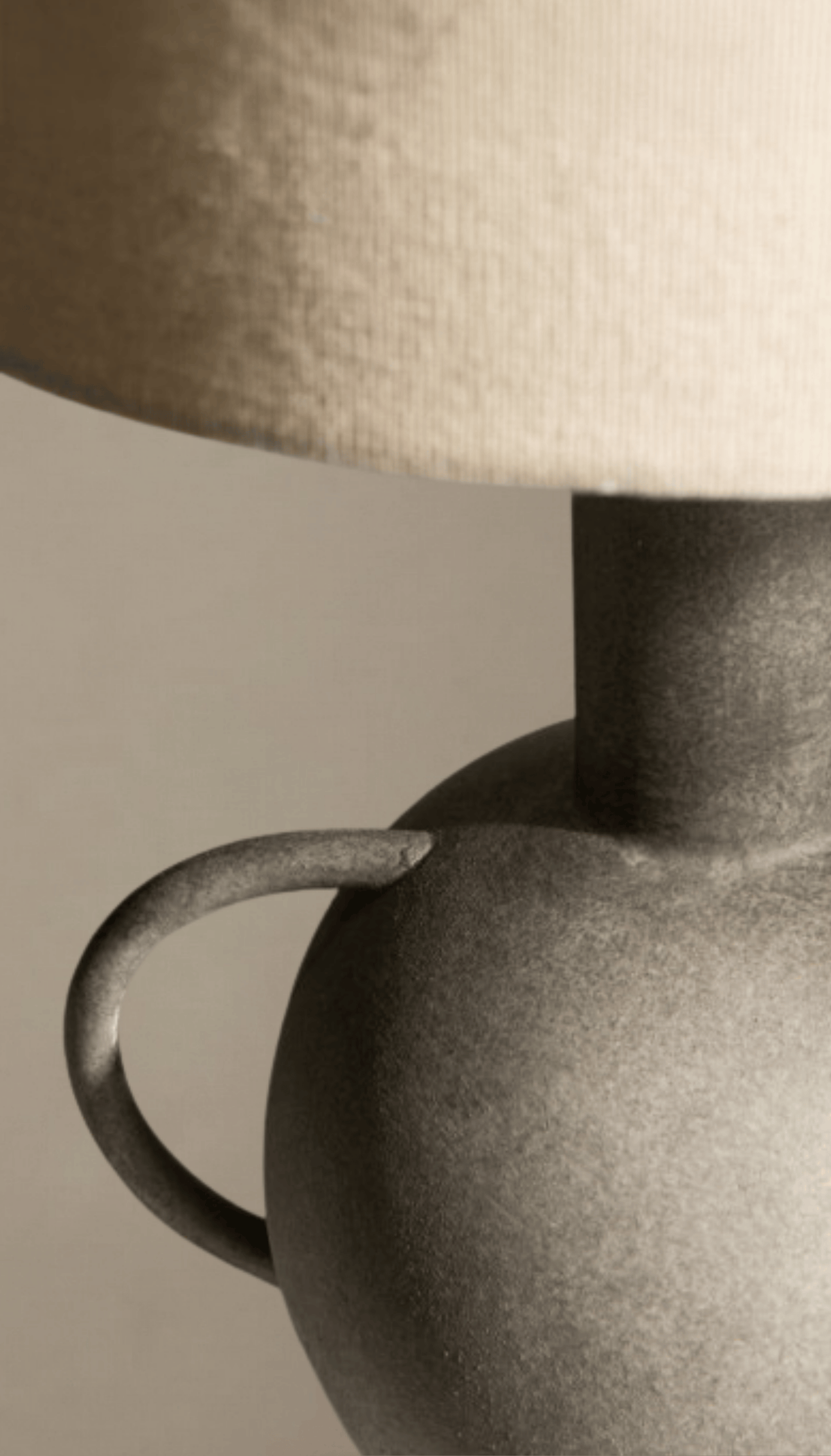 Lámpara de mesa modelo Umbral con base escultural tipo ánfora en gris texturizado efecto piedra, y pantalla cilíndrica de tela de lino color natural.