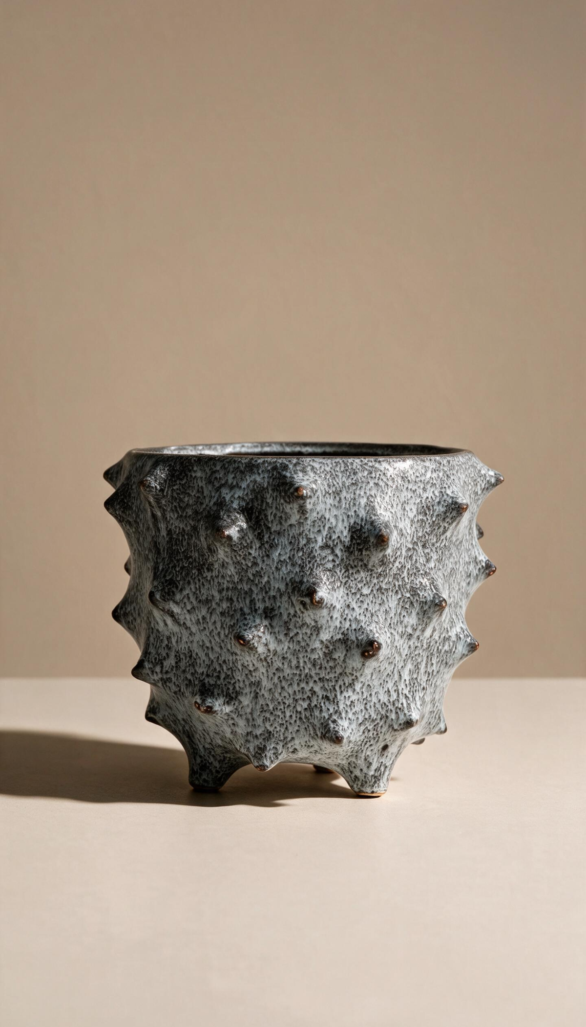 Macetero de gres modelo Brezo en color gris moteado, con diseño rústico de textura con pinchos y base con patas para plantas de interior.
