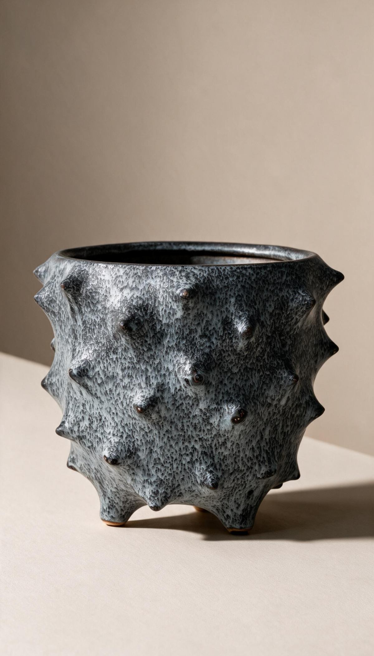 Macetero de gres modelo Brezo en color gris moteado, con diseño rústico de textura con pinchos y base con patas para plantas de interior.