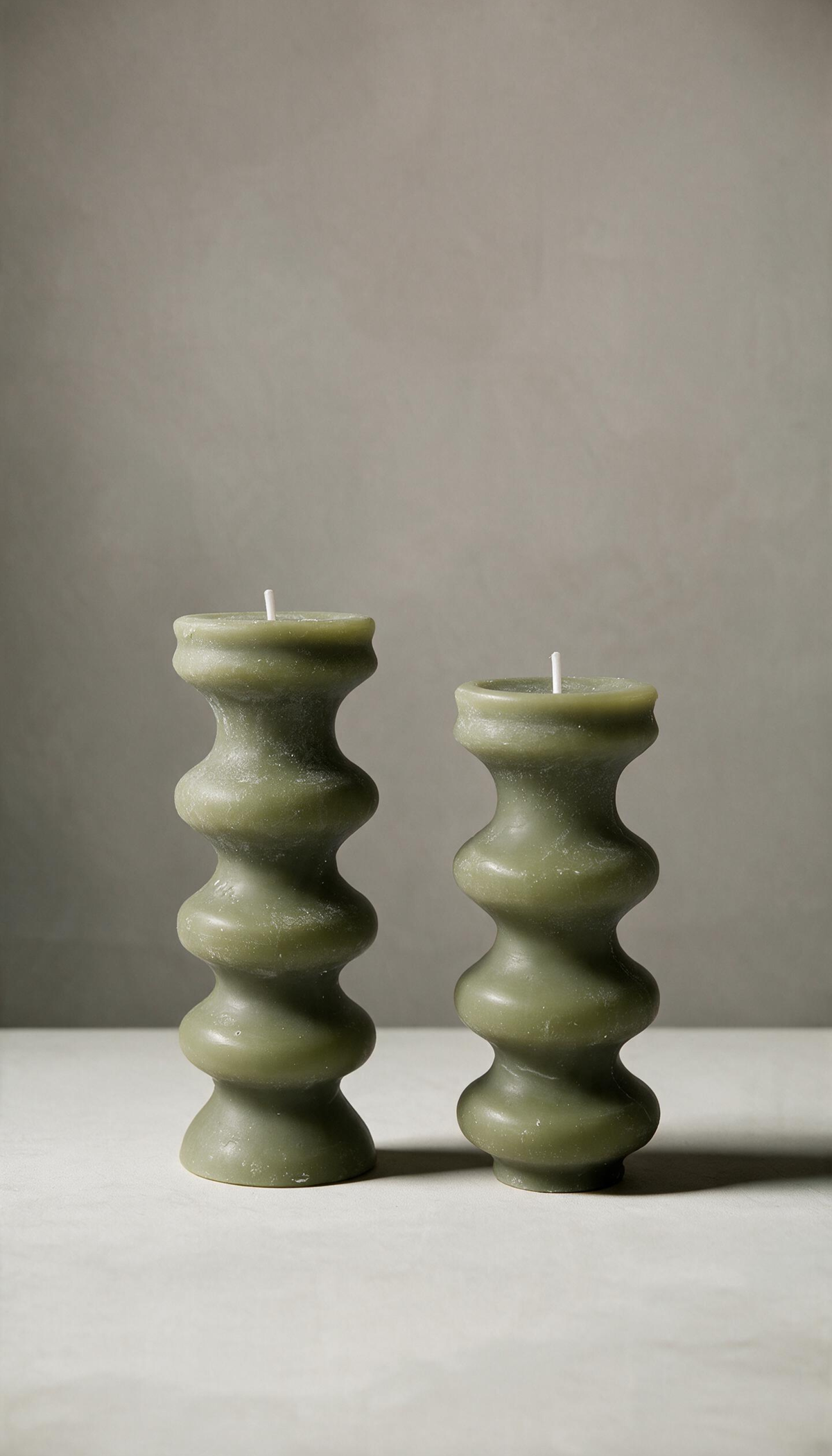 Set de 2 velas decorativas de pilar con diseño torneado escultural de curvas apiladas, en color verde y de diferentes alturas, sobre un fondo neutro.