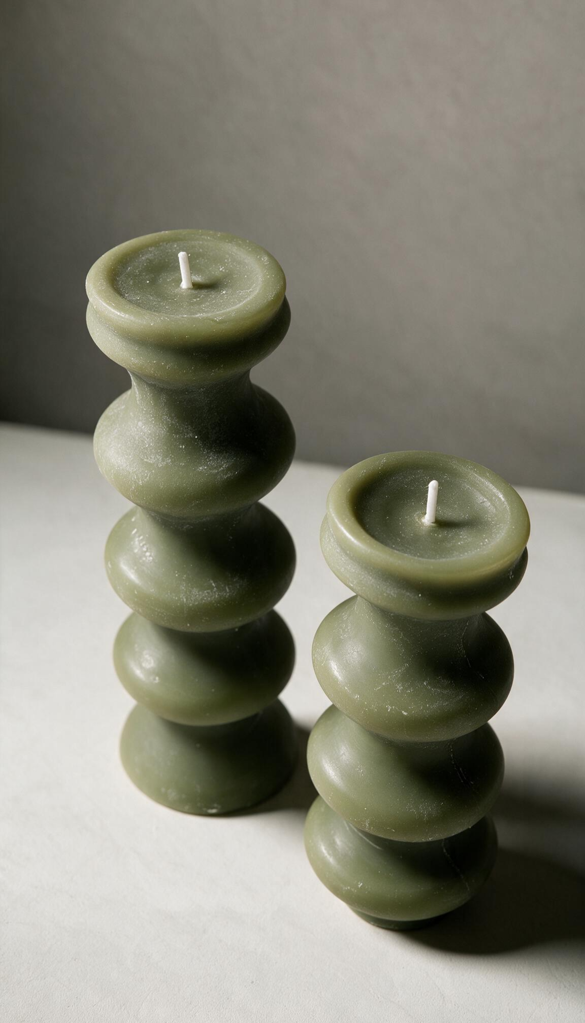 Set de 2 velas decorativas de pilar con diseño torneado escultural de curvas apiladas, en color verde y de diferentes alturas, sobre un fondo neutro.