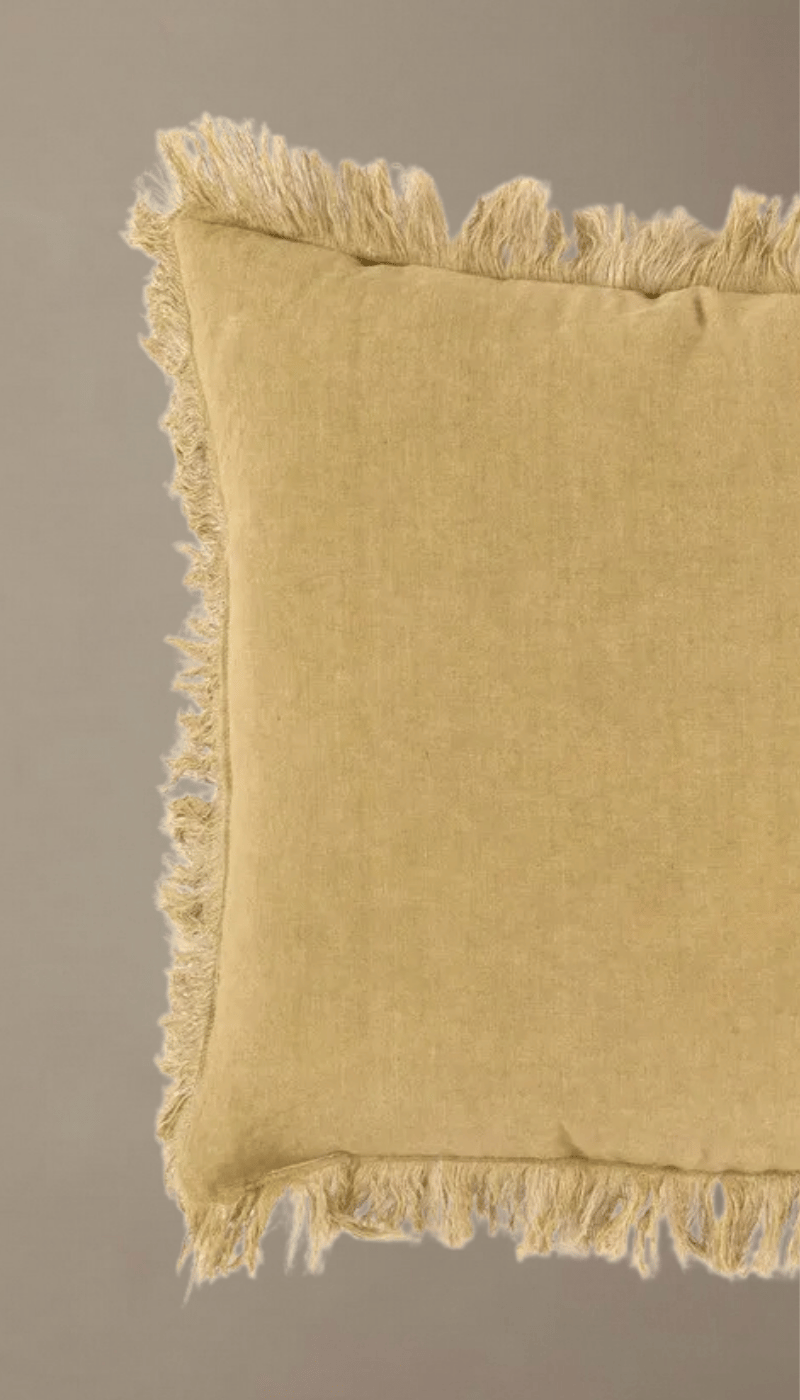 Cojín cuadrado modelo Zahara en color arena o camel, confeccionado en tejido natural estilo lino con bordes decorados con flecos rústicos, apoyado sobre una superficie clara.
