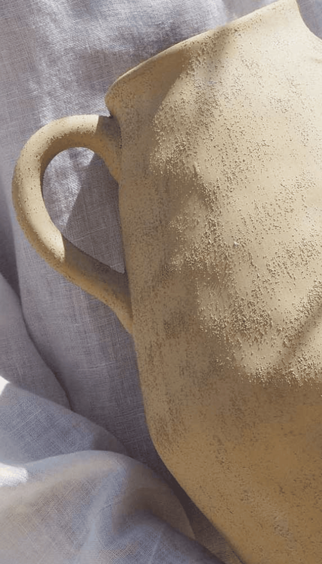 Jarrón de cerámica cruda modelo Mocay en forma de jarra con un asa lateral, color arena claro y textura rugosa, mostrando un marcado estilo rústico y wabi-sabi.